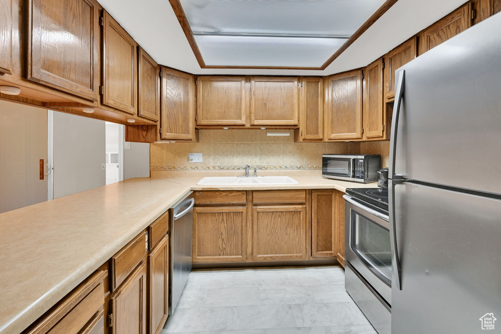 Kitchen featuring stainless steel appliances, light marble finish flooring, a sink, light countertops, and brown cabinetry
