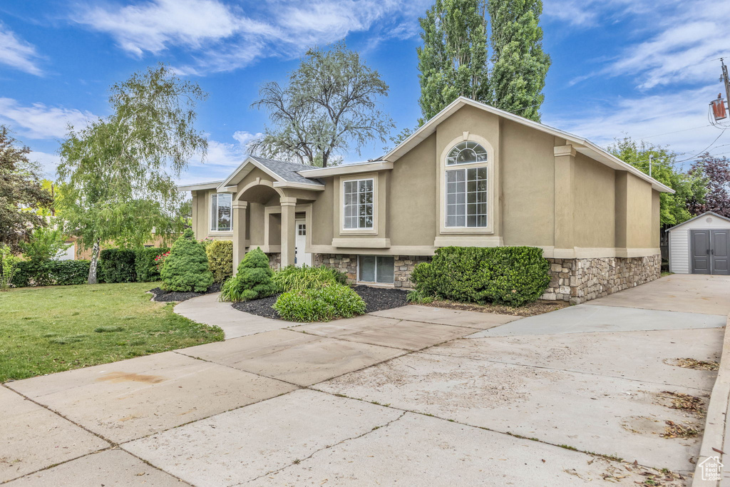 Raised ranch featuring driveway, stucco siding, an outdoor structure, a front lawn, and stone siding