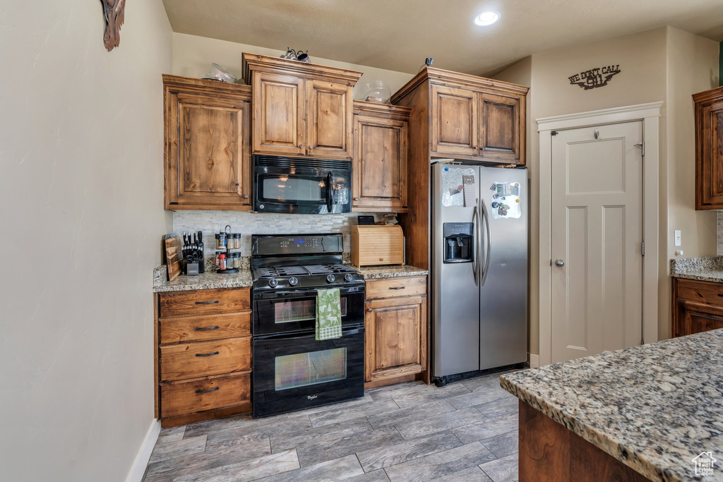 Kitchen with black appliances, decorative backsplash, light stone counters, baseboards, and brown cabinetry