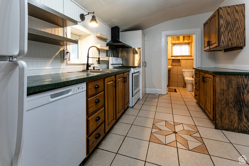 Kitchen featuring white appliances, wall chimney exhaust hood, open shelves, dark countertops, and a sink