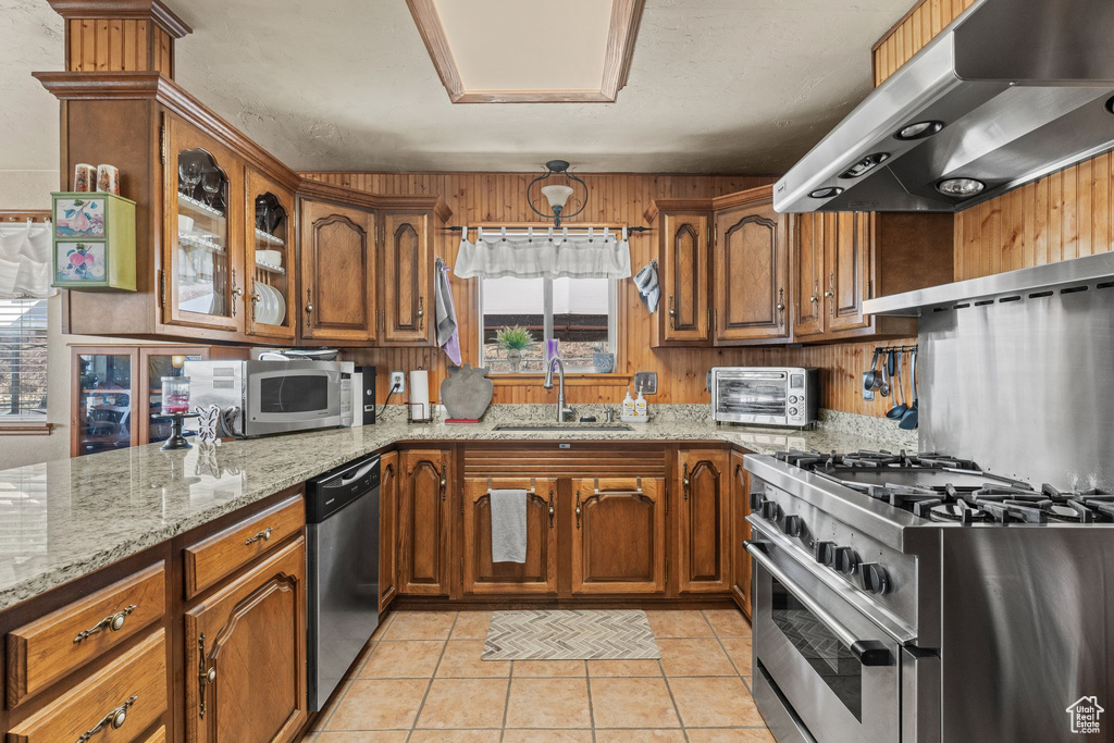 Kitchen featuring range hood, a sink, stainless steel appliances, brown cabinets, and light stone counters