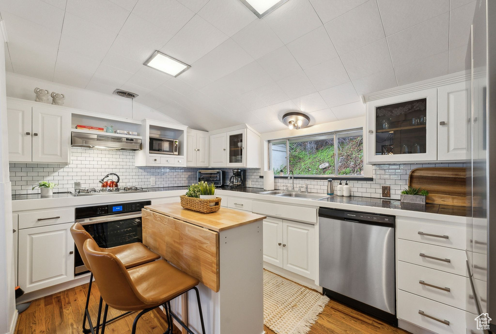Kitchen with appliances with stainless steel finishes, a sink, wood finished floors, white cabinetry, and vaulted ceiling