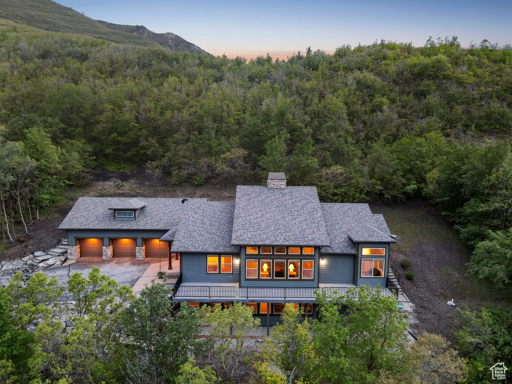 Back of property at dusk with a forest view, a chimney, roof with shingles, and driveway