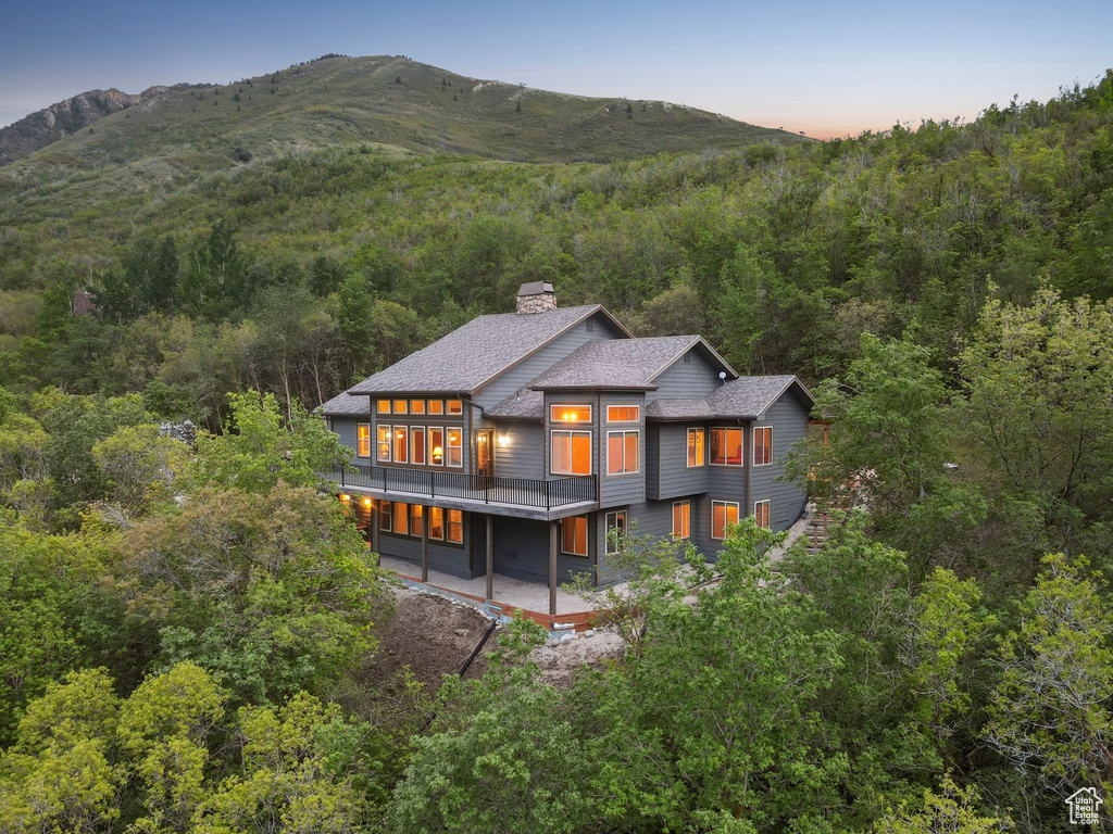 Back of house at dusk with a forest view, a chimney, roof with shingles, a patio area, and a mountain view
