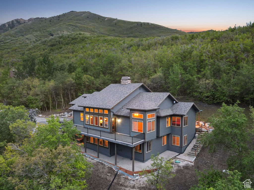 Back of house at dusk featuring a view of trees, a chimney, roof with shingles, and a mountain view
