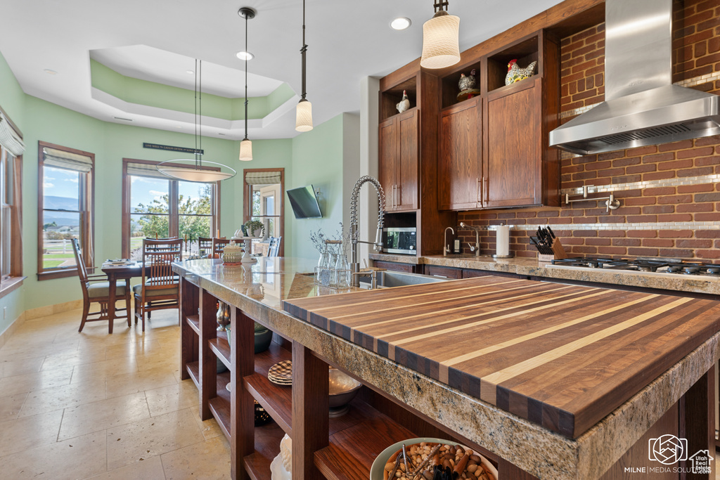 Kitchen with open shelves, wall chimney range hood, backsplash, a raised ceiling, and hanging light fixtures