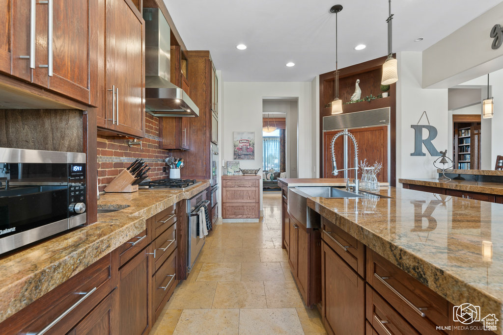 Kitchen featuring wall chimney range hood, appliances with stainless steel finishes, a sink, decorative backsplash, and hanging light fixtures