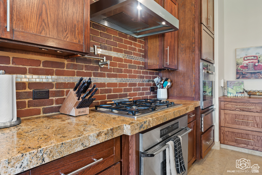 Kitchen with extractor fan, appliances with stainless steel finishes, a warming drawer, tasteful backsplash, and light stone countertops