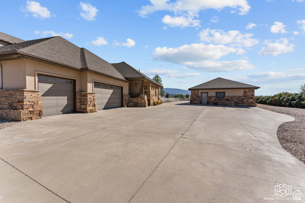 View of side of home with stone siding, a garage, concrete driveway, stucco siding, and roof with shingles