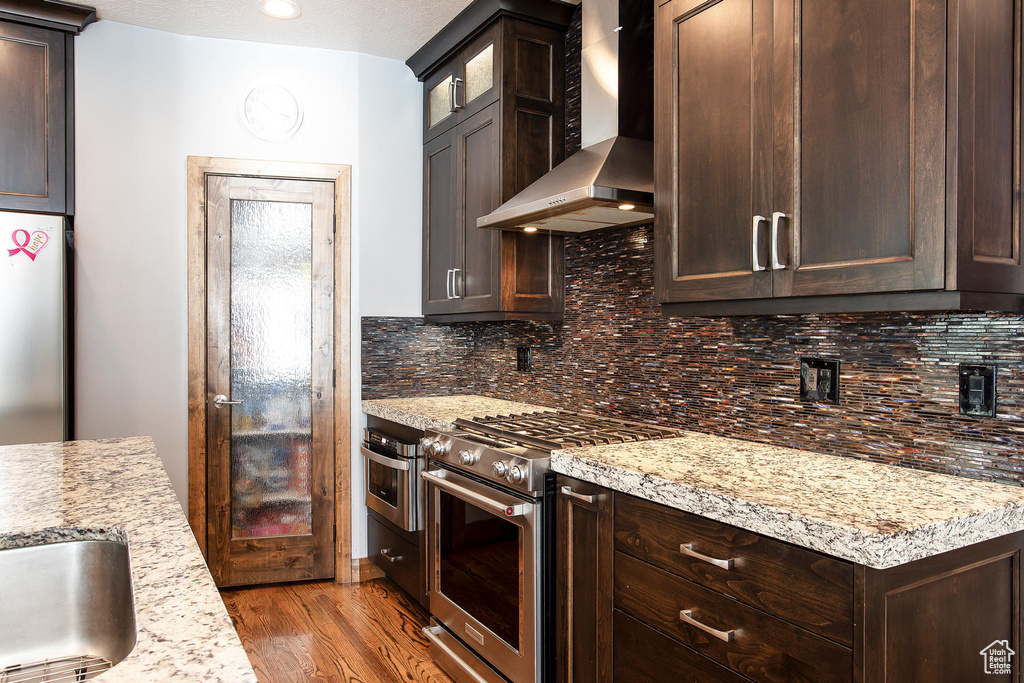 Kitchen featuring wall chimney exhaust hood, appliances with stainless steel finishes, dark brown cabinets, wood finished floors, and decorative backsplash