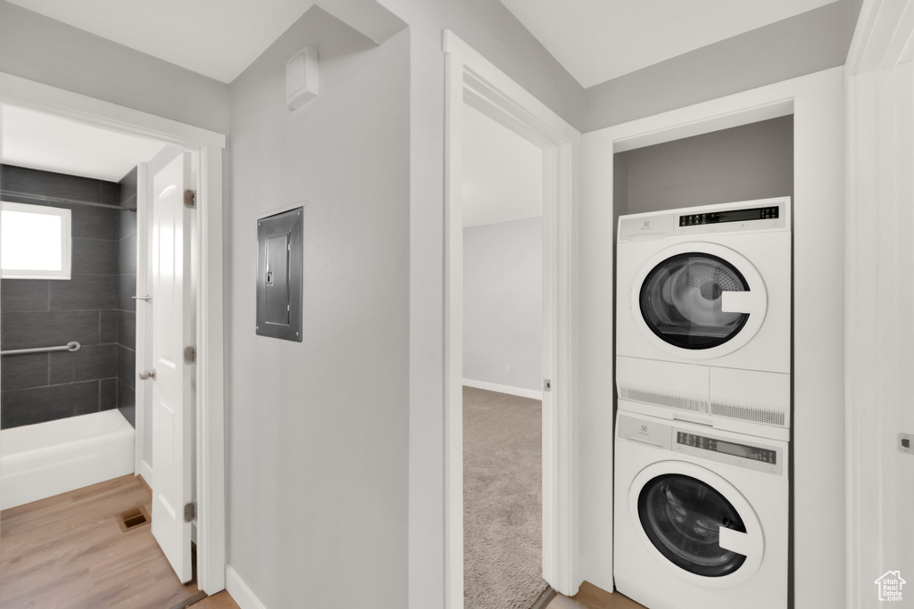 Clothes washing area featuring stacked washing machine and dryer, electric panel, baseboards, and wood finished floors