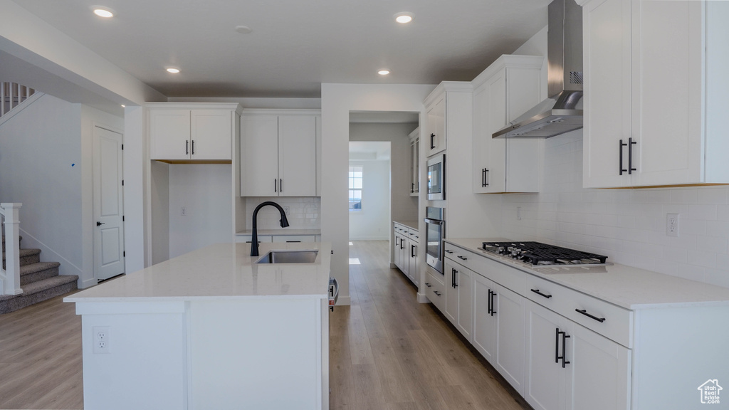 Kitchen featuring wall chimney range hood, stainless steel appliances, light wood finished floors, a kitchen island with sink, and tasteful backsplash