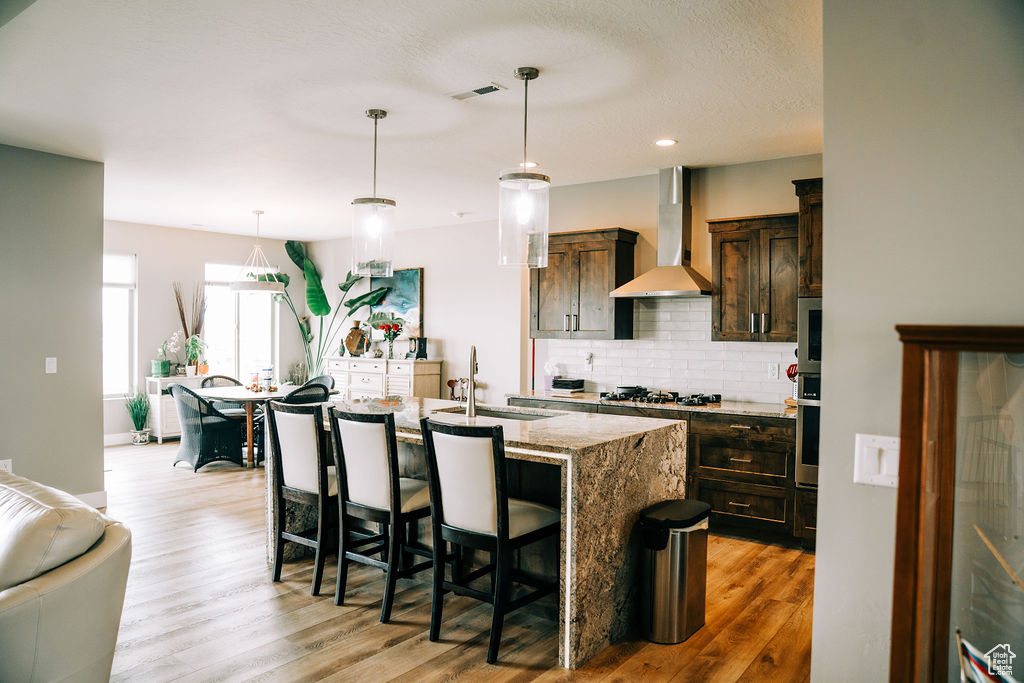 Kitchen featuring wall chimney range hood, a sink, backsplash, light wood-style floors, and light stone counters