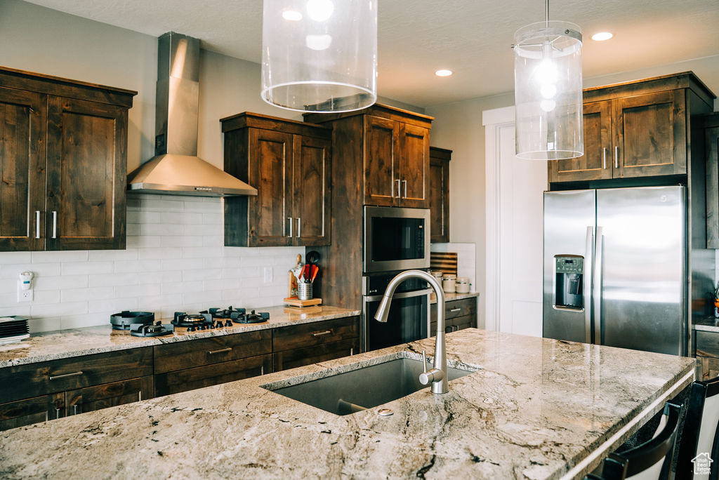 Kitchen featuring wall chimney range hood, appliances with stainless steel finishes, a sink, decorative backsplash, and light stone counters