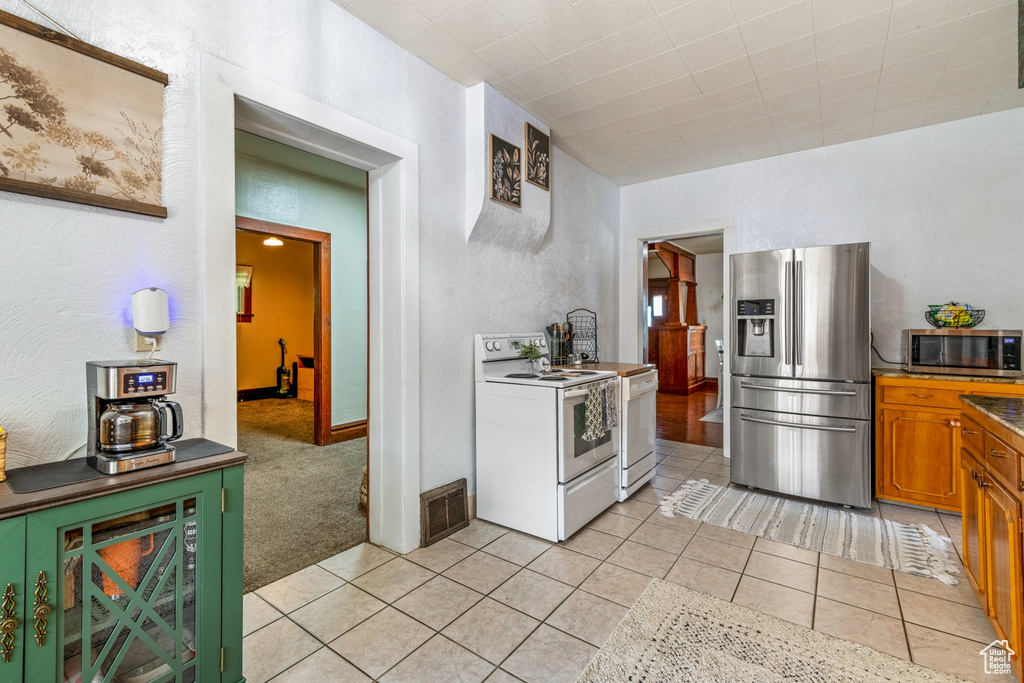 Kitchen with appliances with stainless steel finishes, brown cabinetry, a textured wall, light tile patterned flooring, and light colored carpet