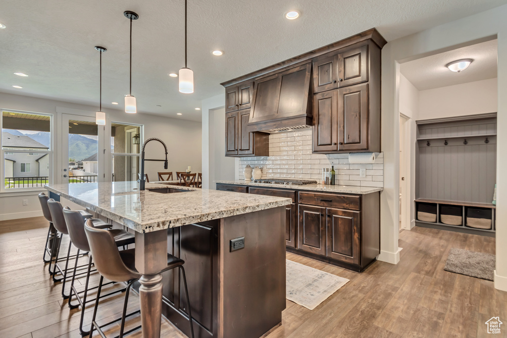 Kitchen with custom exhaust hood, a sink, light wood-style floors, dark brown cabinets, and backsplash