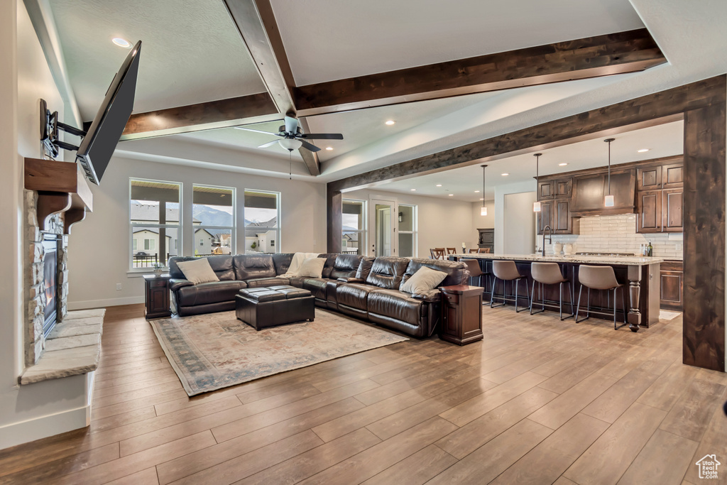 Living room with a ceiling fan, beamed ceiling, light wood finished floors, baseboards, and a stone fireplace