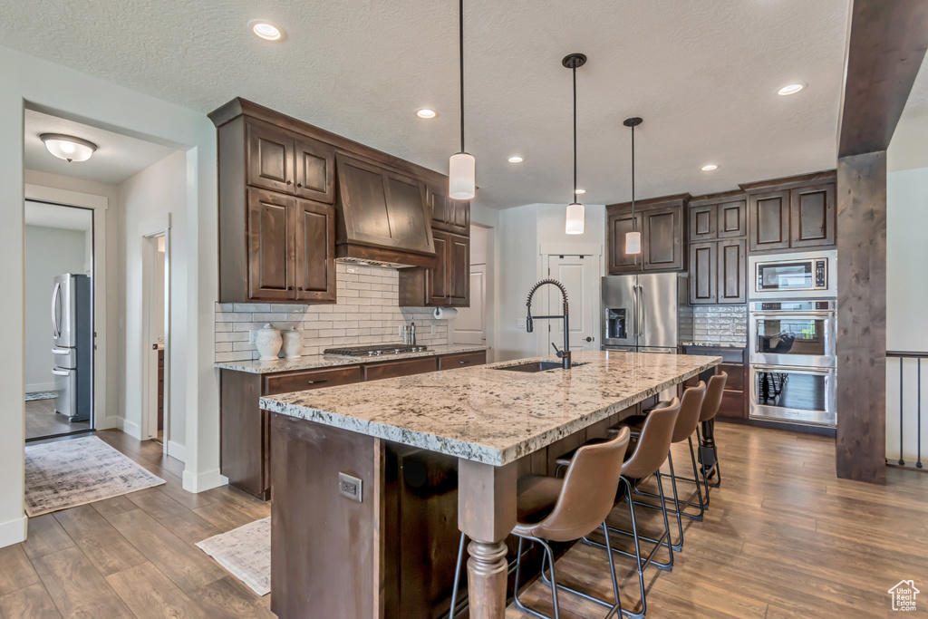 Kitchen featuring appliances with stainless steel finishes, premium range hood, dark brown cabinets, tasteful backsplash, and a sink