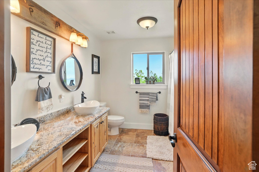 Bathroom featuring toilet, double vanity, stone finish floors, baseboards, and a shower with curtain