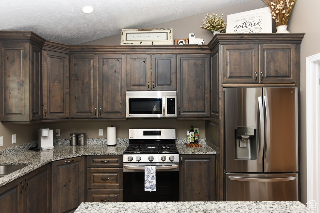 Kitchen with stainless steel appliances, dark brown cabinets, lofted ceiling, and light stone counters