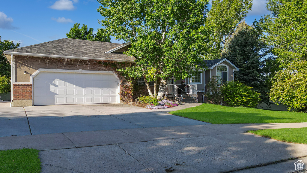 View of property hidden behind natural elements featuring driveway, a front yard, an attached garage, roof with shingles, and brick siding