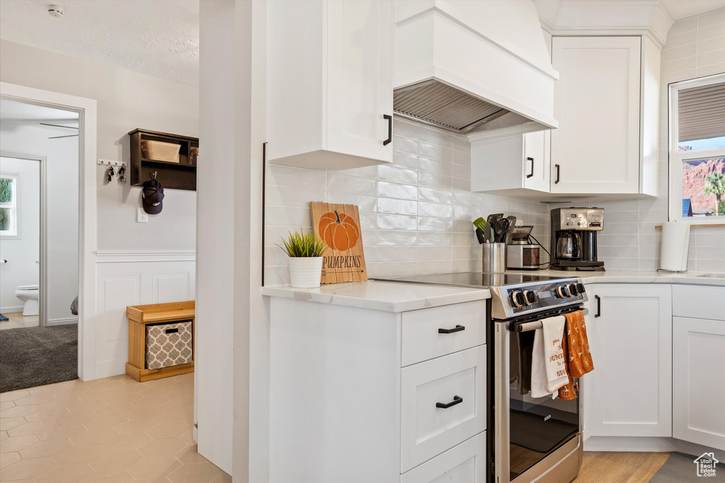 Kitchen with custom exhaust hood, stainless steel electric range oven, white cabinets, and backsplash