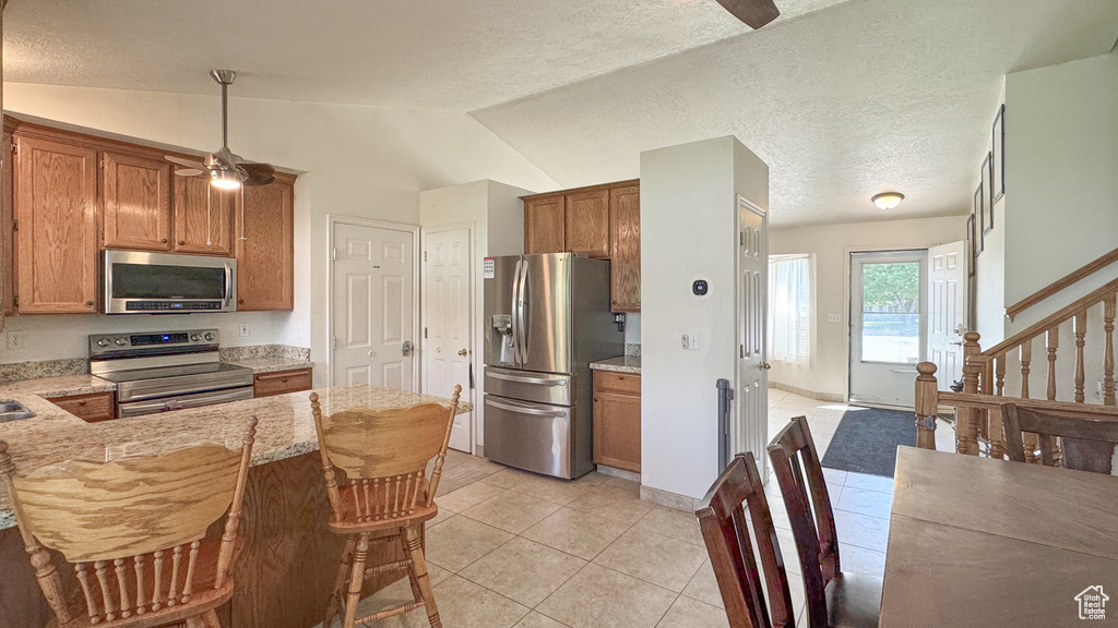 Kitchen featuring stainless steel appliances, brown cabinetry, ceiling fan, light tile patterned floors, and vaulted ceiling