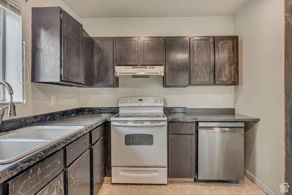 Kitchen featuring stainless steel dishwasher, white electric stove, under cabinet range hood, dark countertops, and a sink