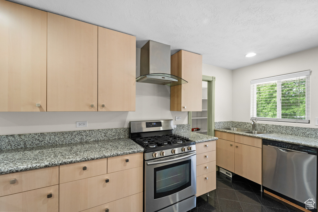 Kitchen with appliances with stainless steel finishes, wall chimney range hood, light brown cabinetry, a sink, and granite floors