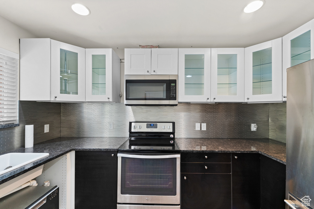 Kitchen featuring stainless steel appliances, white cabinetry, decorative backsplash, dark stone countertops, and glass insert cabinets