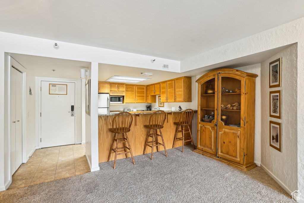 Kitchen with stainless steel microwave, freestanding refrigerator, light carpet, brown cabinetry, and a breakfast bar area