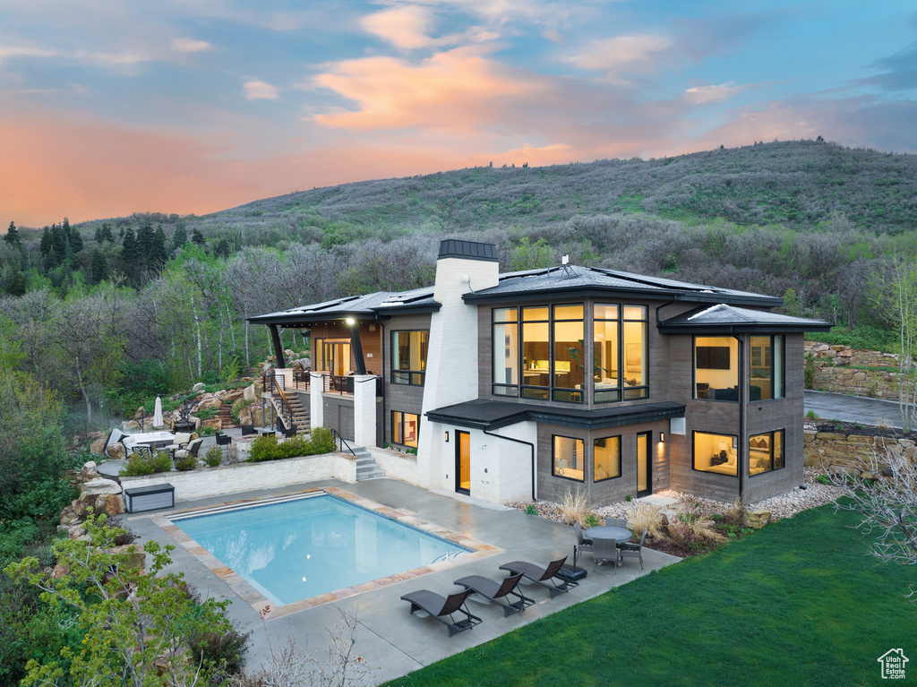 Back of house at dusk with stairs, a balcony, an outdoor pool, a patio area, and a wooded view