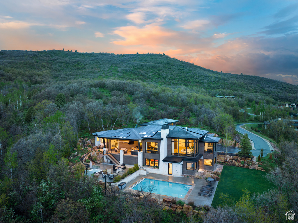 Back of house at dusk featuring stairs, a wooded view, an outdoor pool, a patio area, and a jacuzzi