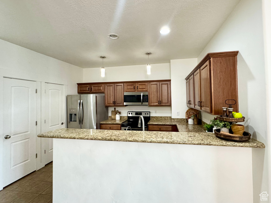 Kitchen featuring stainless steel appliances, a peninsula, brown cabinetry, light stone countertops, and pendant lighting