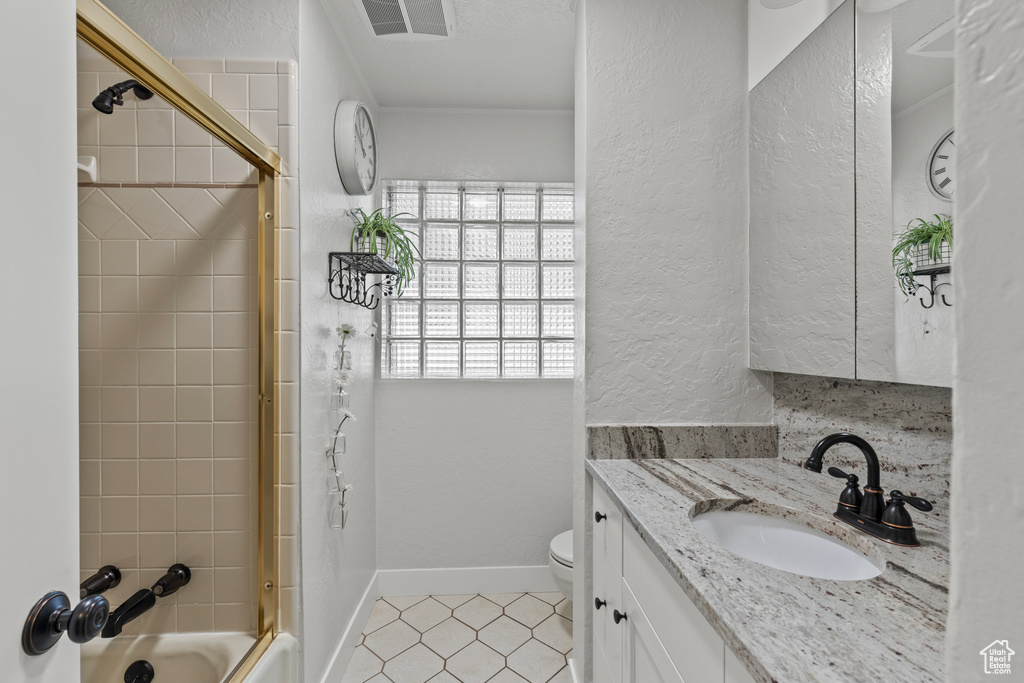 Bathroom featuring tile patterned floors, toilet, vanity, a textured wall, and bath / shower combo with glass door
