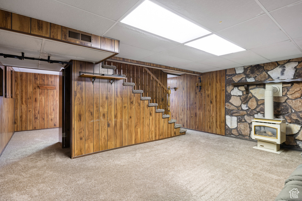 Finished basement featuring a drop ceiling, a wood stove, carpet floors, stairs, and wood walls