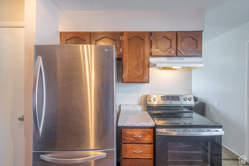 Kitchen featuring stainless steel appliances, under cabinet range hood, brown cabinetry, light countertops, and a textured ceiling