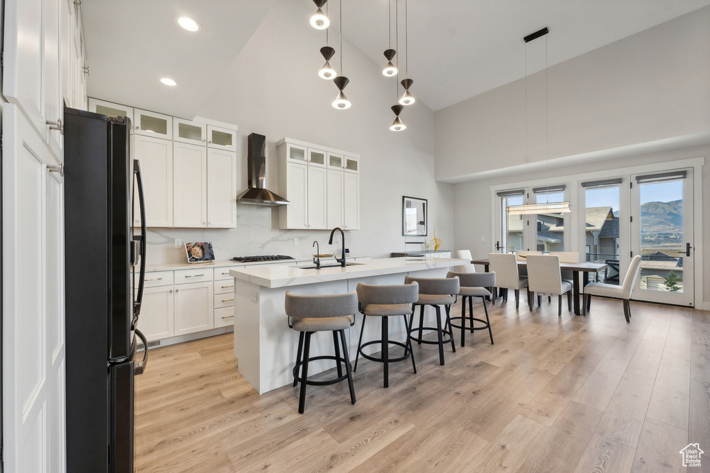 Kitchen with freestanding refrigerator, wall chimney range hood, high vaulted ceiling, light wood-style flooring, and a kitchen island with sink