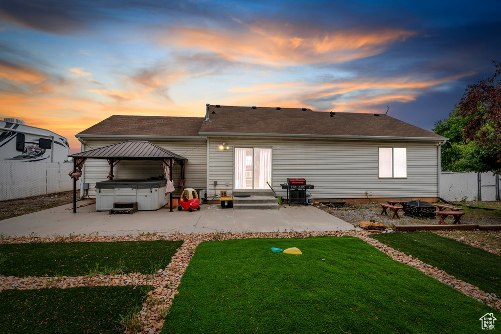 Back of house at dusk with a hot tub, an outdoor fire pit, a patio area, roof with shingles, and a gazebo