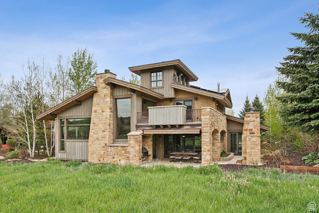 Back of house featuring a balcony, a chimney, a lawn, stone siding, and a patio