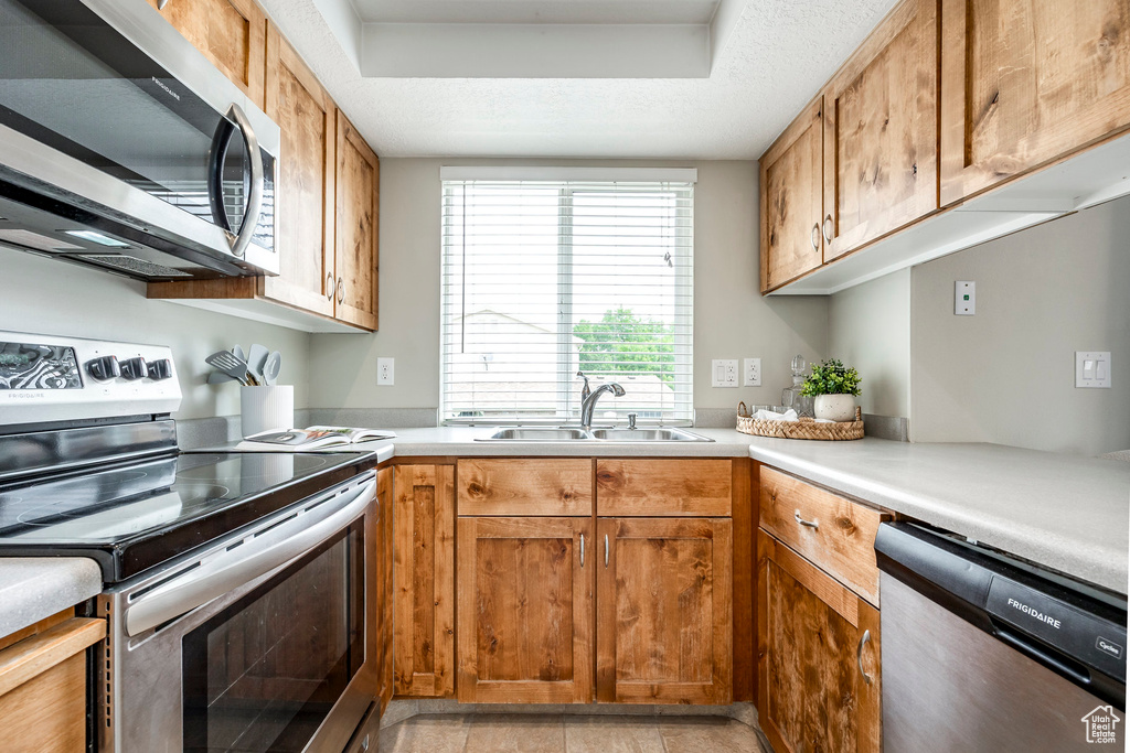 Kitchen featuring appliances with stainless steel finishes, a sink, light countertops, a tray ceiling, and brown cabinetry