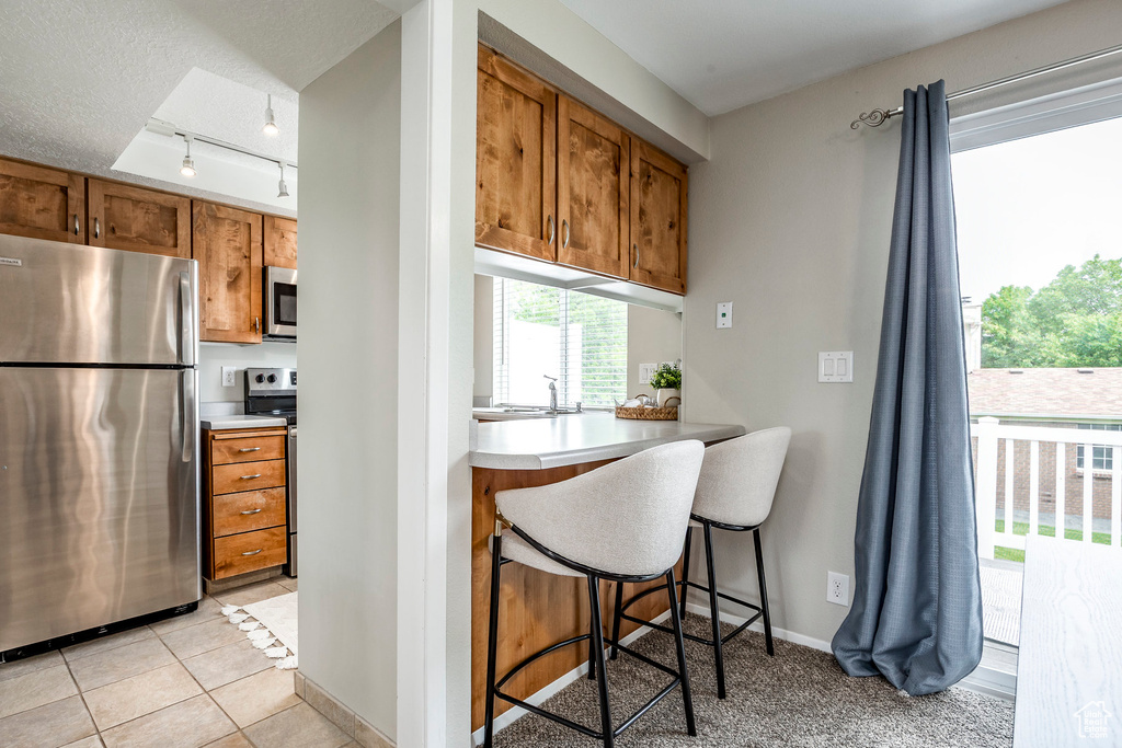 Kitchen with stainless steel appliances, brown cabinets, rail lighting, light countertops, and a kitchen bar