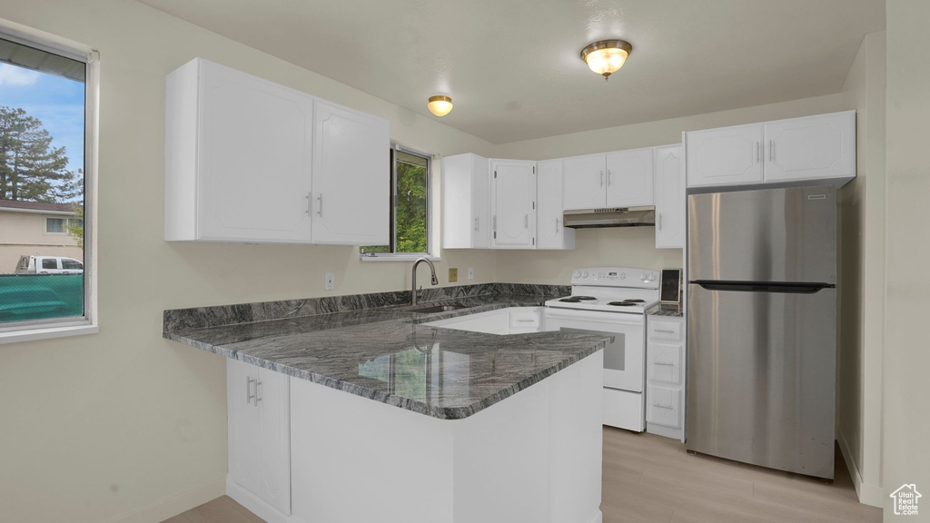 Kitchen with freestanding refrigerator, white electric range oven, under cabinet range hood, dark stone countertops, and a peninsula