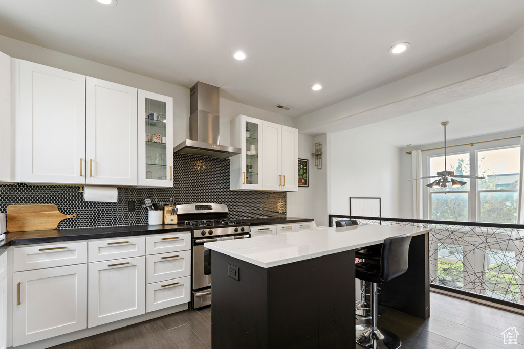 Kitchen with wall chimney range hood, stainless steel gas stove, dark wood-style flooring, backsplash, and white cabinetry
