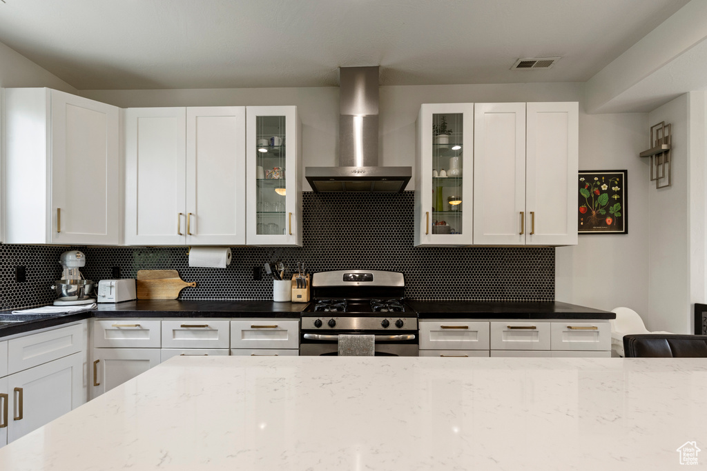 Kitchen featuring wall chimney exhaust hood, stainless steel gas stove, tasteful backsplash, dark stone counters, and white cabinets