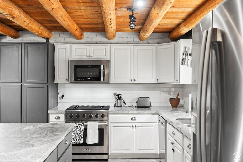 Kitchen featuring appliances with stainless steel finishes, decorative backsplash, wood ceiling with exposed beams, white cabinetry, and light stone countertops