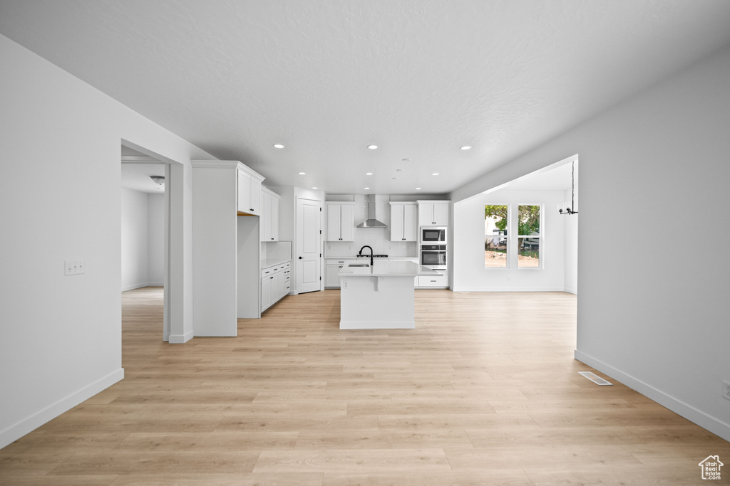 Kitchen with wall chimney range hood, built in microwave, white cabinets, a kitchen island with sink, and wall oven
