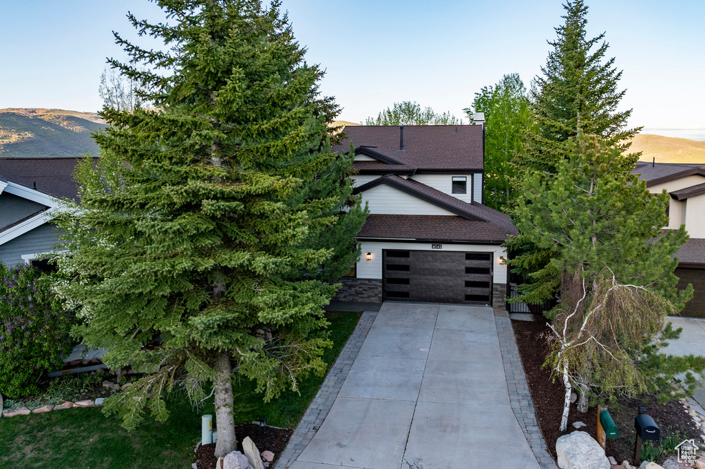 View of property hidden behind natural elements with driveway, stone siding, and roof with shingles
