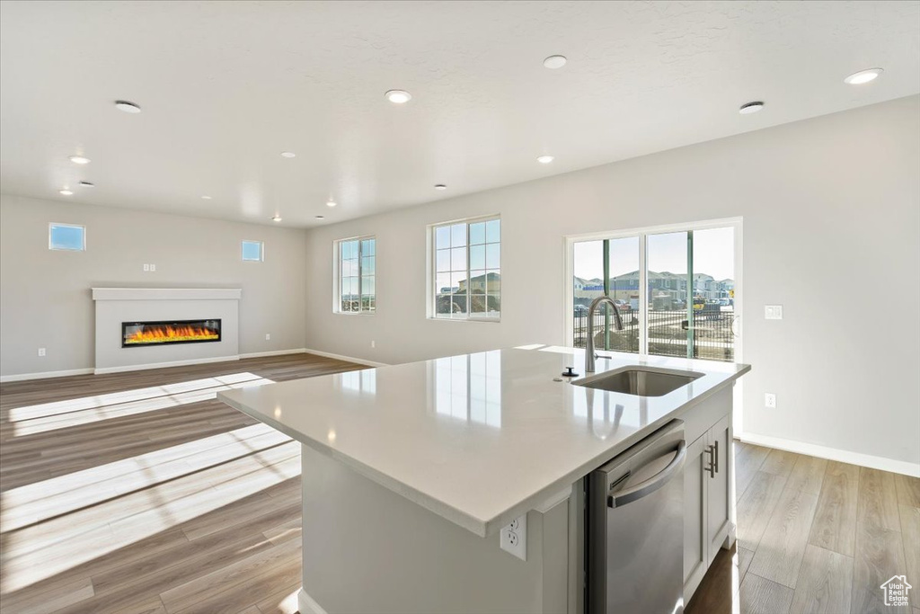 Kitchen featuring stainless steel dishwasher, a sink, light wood-style flooring, a glass covered fireplace, and a center island with sink