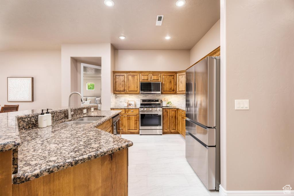 Kitchen with stainless steel appliances, a sink, brown cabinets, decorative backsplash, and light stone counters